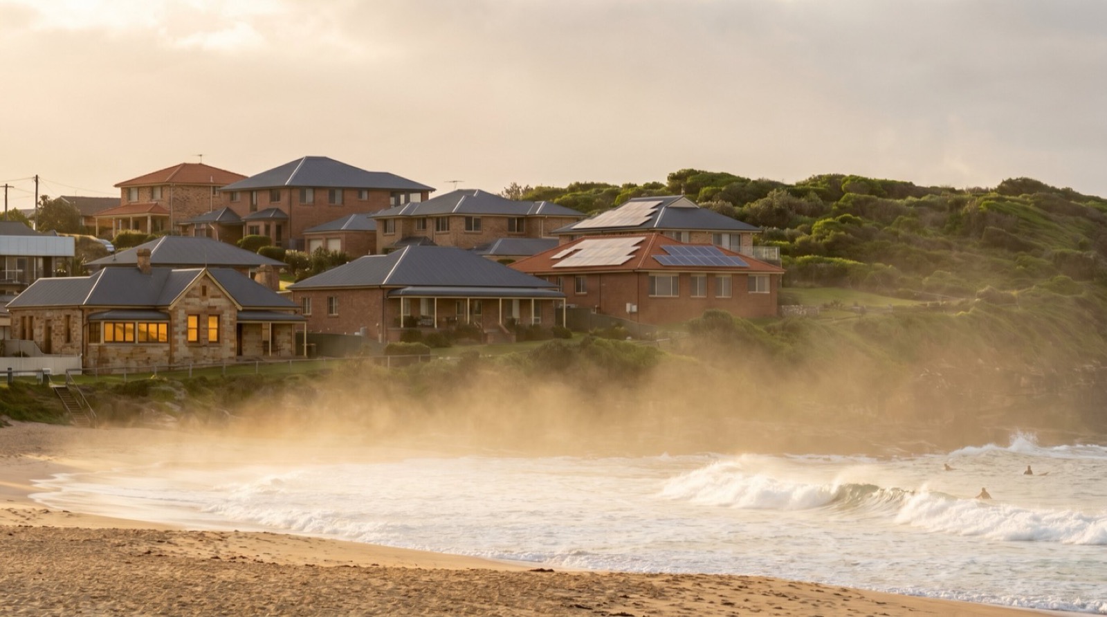 Golden-hour view along a Newcastle surf beach, salt spray hazing up off the breaking waves toward established brick-and-Colorbond houses on the low headland behind, a couple of them with rooftop solar panels